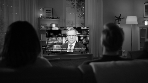 kaksi henkilöä katsoo televisiota, jossa näkyy uutistenlukija ja tekstitykset