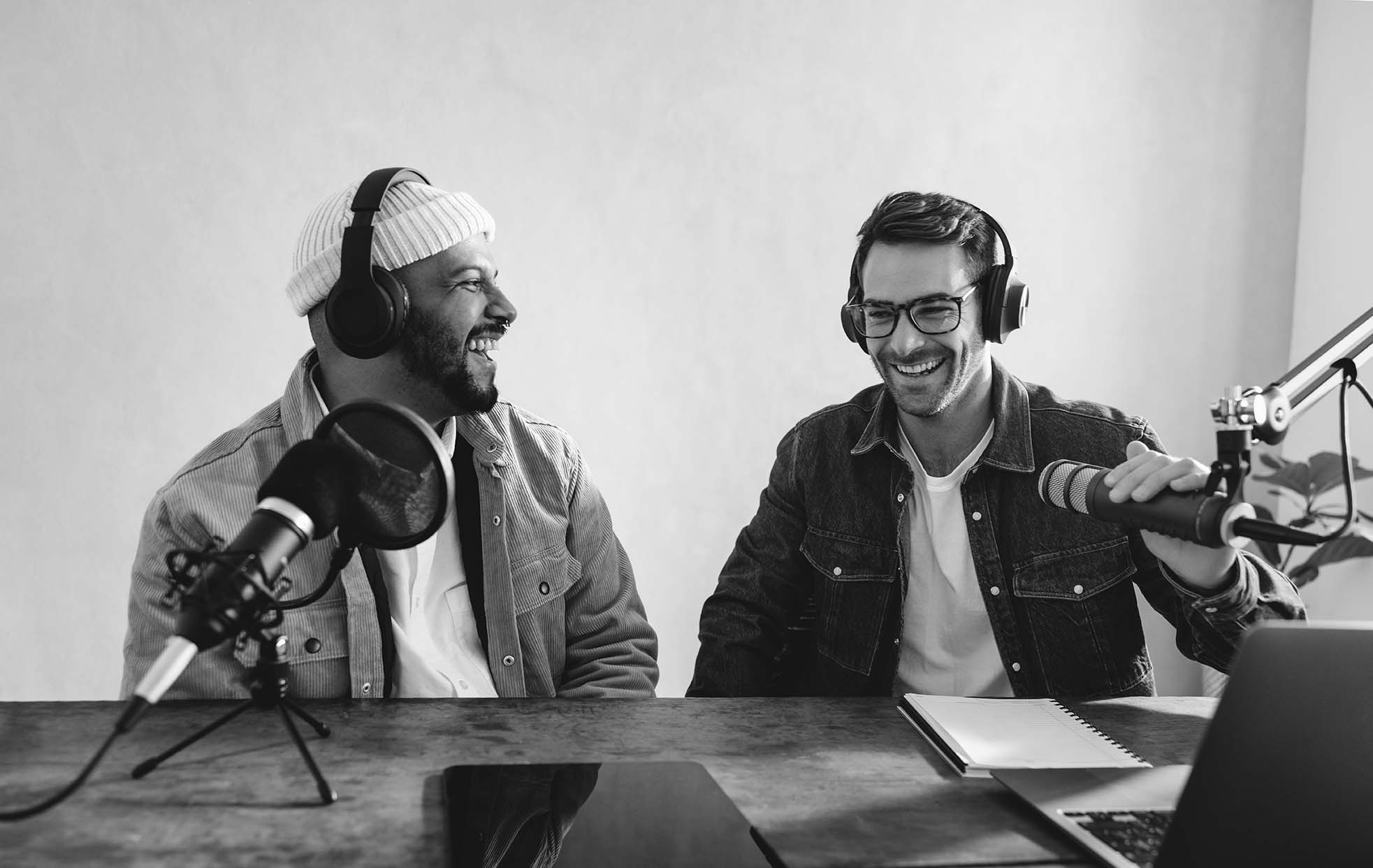 Kaksi nauravaa mieltä kuulokkeet päässään äänitysmikrofonien kanssa. He nauhoittavat podcast-jaksoa. Jakson litteraatti löytyy hakukoneilla ja parantaa podcastin löydettävyyttä.